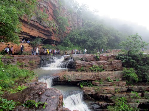 Tourists walking along the rocky cliffs and cascading streams of Mallela Theertham Waterfalls, surrounded by lush greenery and mist in the Nallamala forest