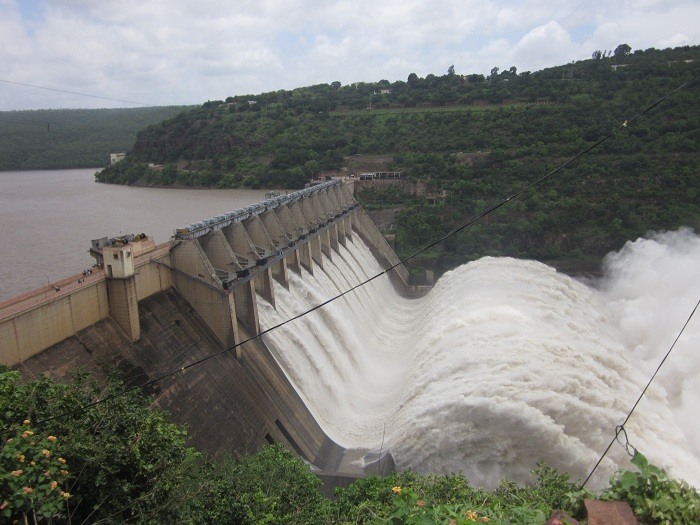 Nagarjunasagar Dam releasing water through its spillway gates, surrounded by lush green hills and cloudy skies near Nallamala Forest & Tiger Reserve