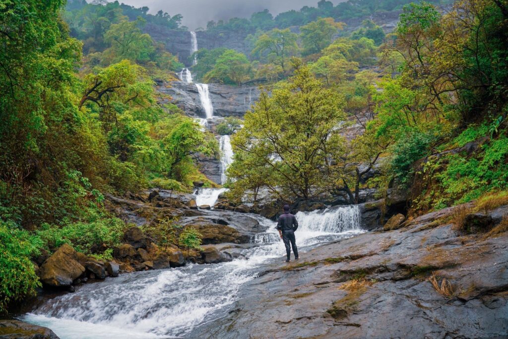 Trekking enthusiast exploring a multi-tiered waterfall surrounded by dense forest and rocky terrain, with cascading streams and vibrant greenery creating a scenic adventure destination.